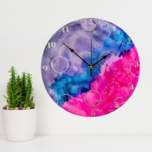 A round wall clock with a vibrant alcohol ink design in purple, blue, and pink, featuring silver Arabic numerals, silver "bubbles" and black hands.