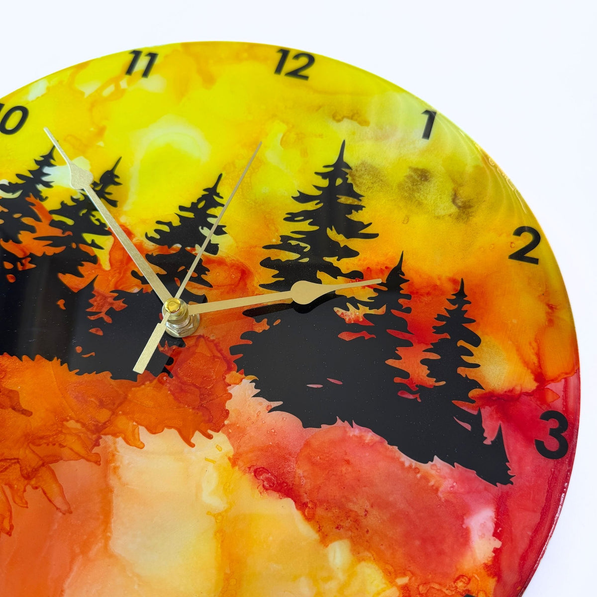 A round wall clock with a vibrant alcohol ink landscape design featuring black pine tree silhouettes over a gradient of yellow, orange, and red, with black Arabic numerals and gold hands.