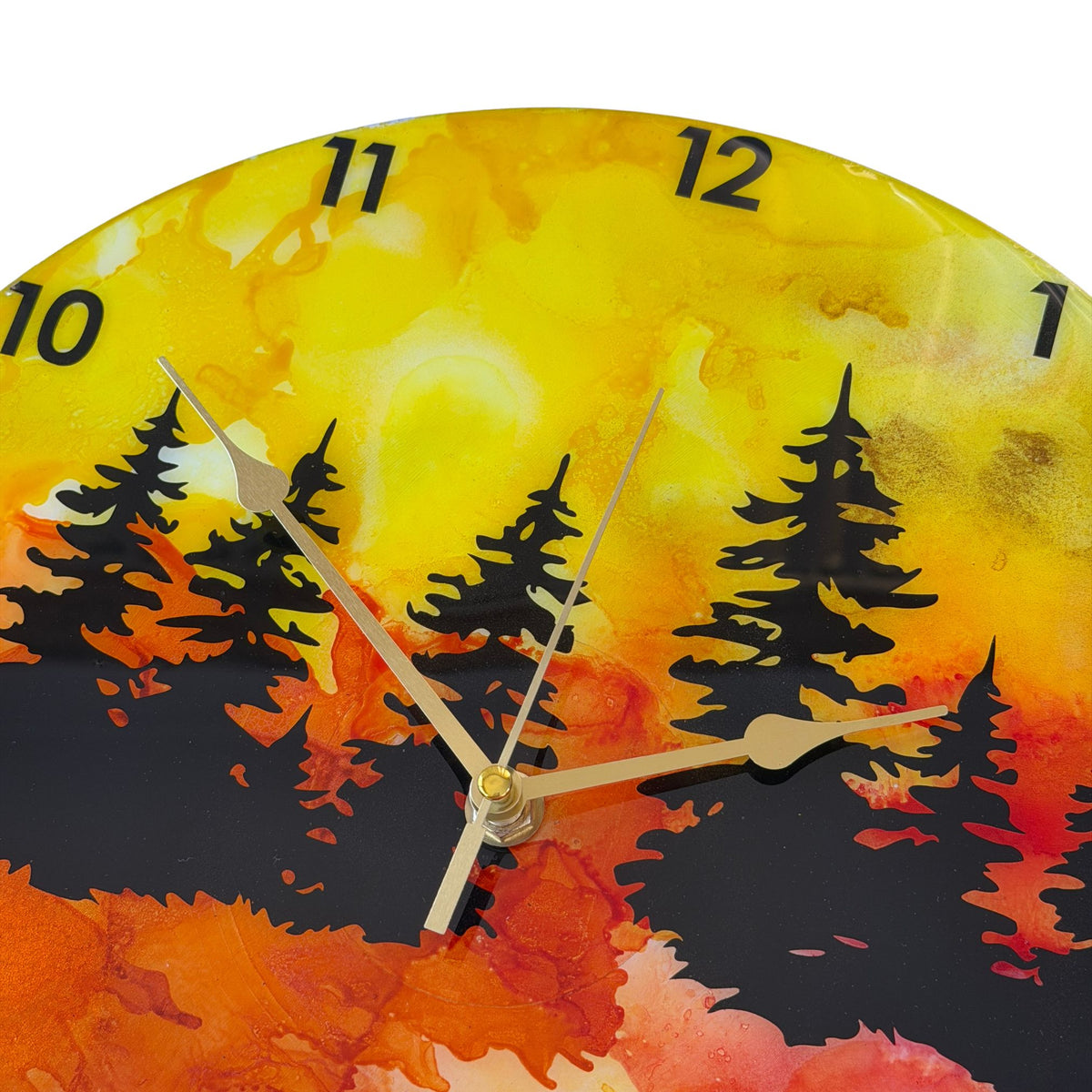 A round wall clock with a vibrant alcohol ink landscape design featuring black pine tree silhouettes over a gradient of yellow, orange, and red, with black Arabic numerals and gold hands.