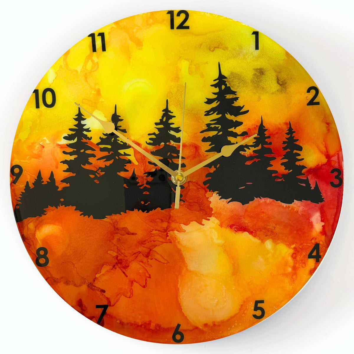 A round wall clock with a vibrant alcohol ink landscape design featuring black pine tree silhouettes over a gradient of yellow, orange, and red, with black Arabic numerals and gold hands.