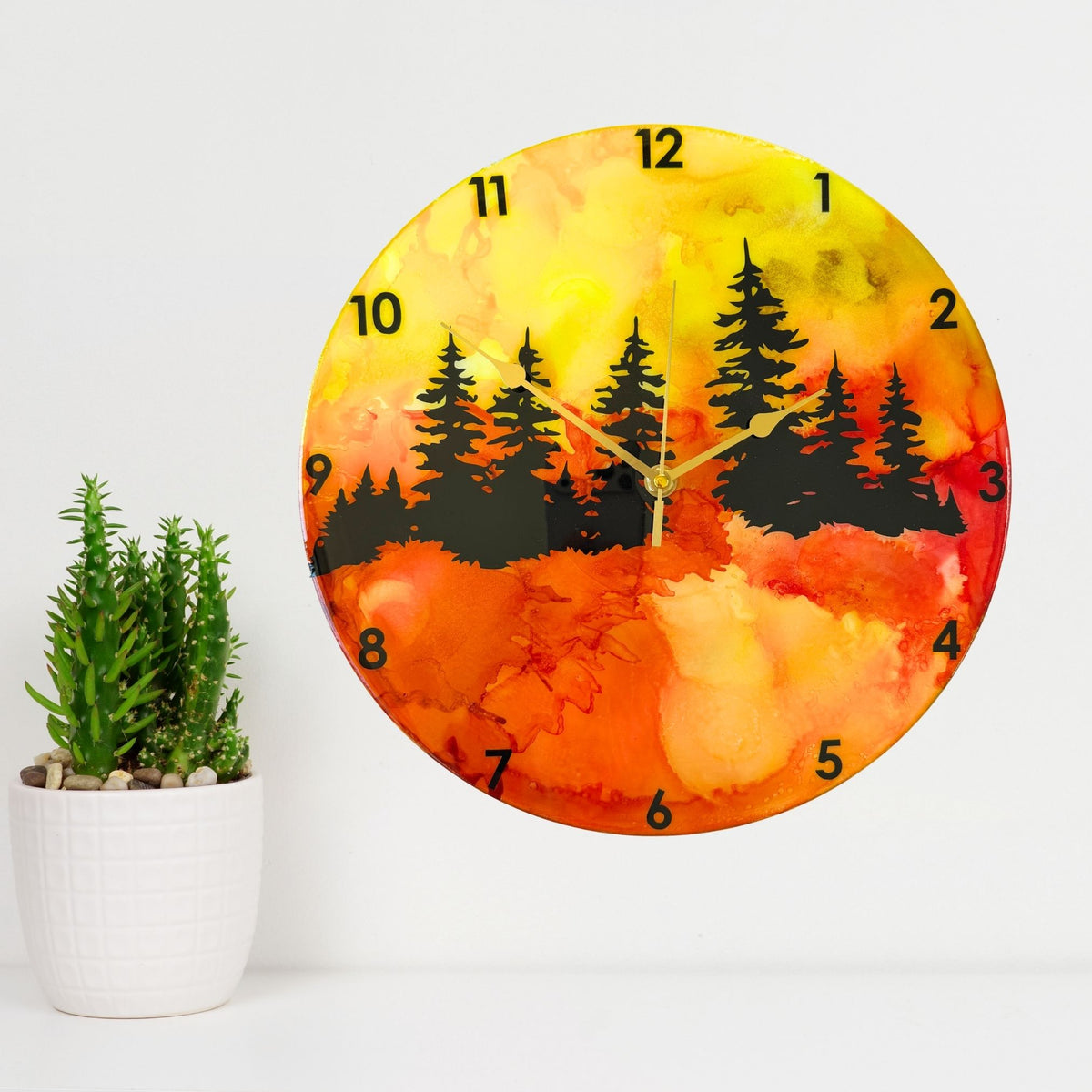 A round wall clock with a vibrant alcohol ink landscape design featuring black pine tree silhouettes over a gradient of yellow, orange, and red, with black Arabic numerals and gold hands.