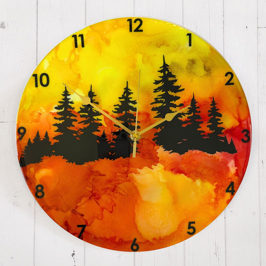 A round wall clock with a vibrant alcohol ink landscape design featuring black pine tree silhouettes over a gradient of yellow, orange, and red, with black Arabic numerals and gold hands.