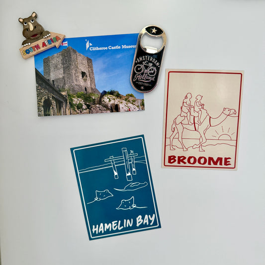 A set of travel-themed magnets or stickers displayed on a white refrigerator, including a Clifton Castle Museum magnet, an Amsterdam Bike Festival bottle opener magnet, a Broome illustration magnet, and a Hamelin Bay magnet.