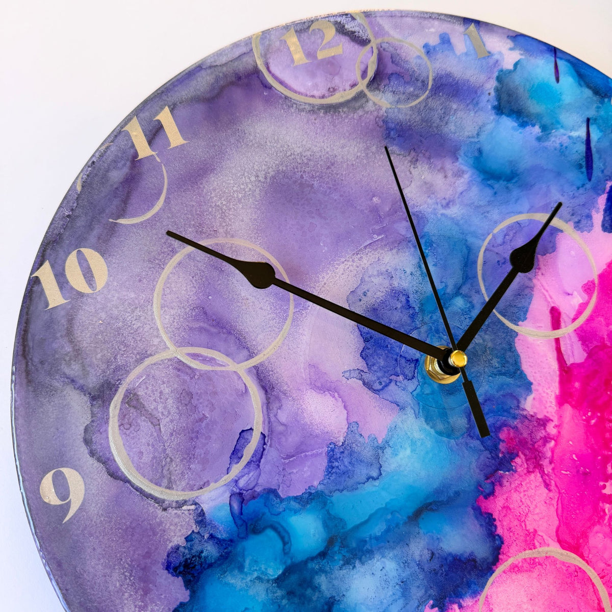 A round watercolor-style wall clock with a purple, blue, and pink abstract design, black hands, and gold Arabic numerals on a white background.