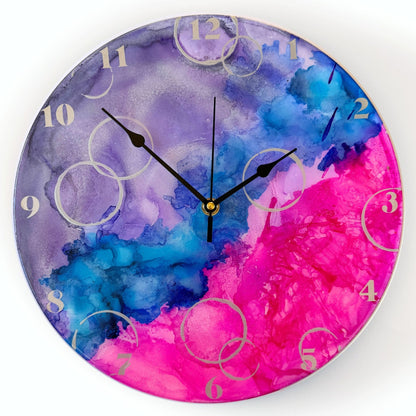 Round wall clock with a vibrant design in purple, blue, and pink, featuring black hands and light silver Arabic numerals.