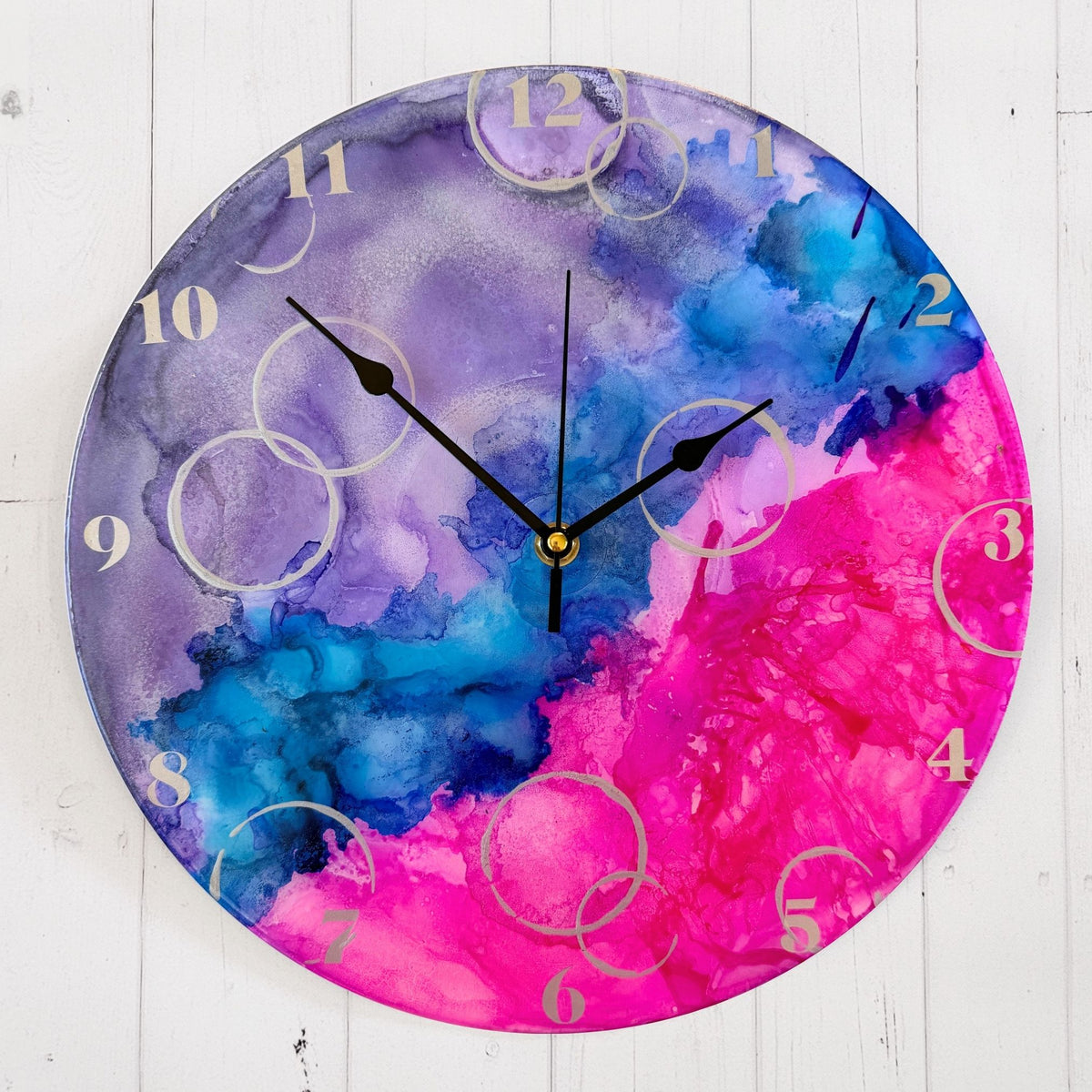 A round wall clock with a vibrant alcohol ink design in purple, blue, and pink, featuring silver Arabic numerals, silver "bubbles" and black hands.
