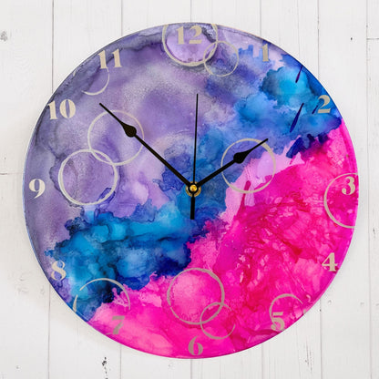 A round wall clock with a vibrant alcohol ink design in purple, blue, and pink, featuring silver Arabic numerals, silver "bubbles" and black hands.