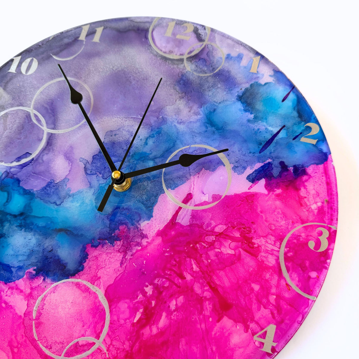 A round wall clock with a vibrant alcohol ink design in purple, blue, and pink, featuring silver Arabic numerals, silver "bubbles" and black hands.