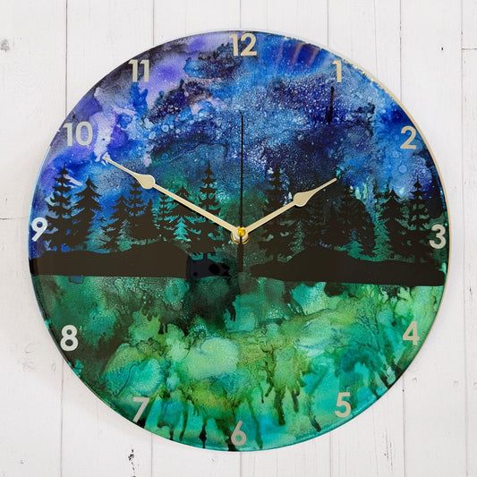 Round wall clock with a scenic forest landscape print in blue, green, and black, featuring silver hands and white Arabic numerals.