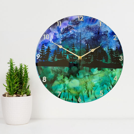 Round wall clock with a scenic forest landscape print in blue, green, and black, featuring silver hands and white Arabic numerals.