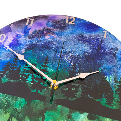 Round wall clock with a scenic forest landscape print in blue, green, and black, featuring silver hands and white Arabic numerals.