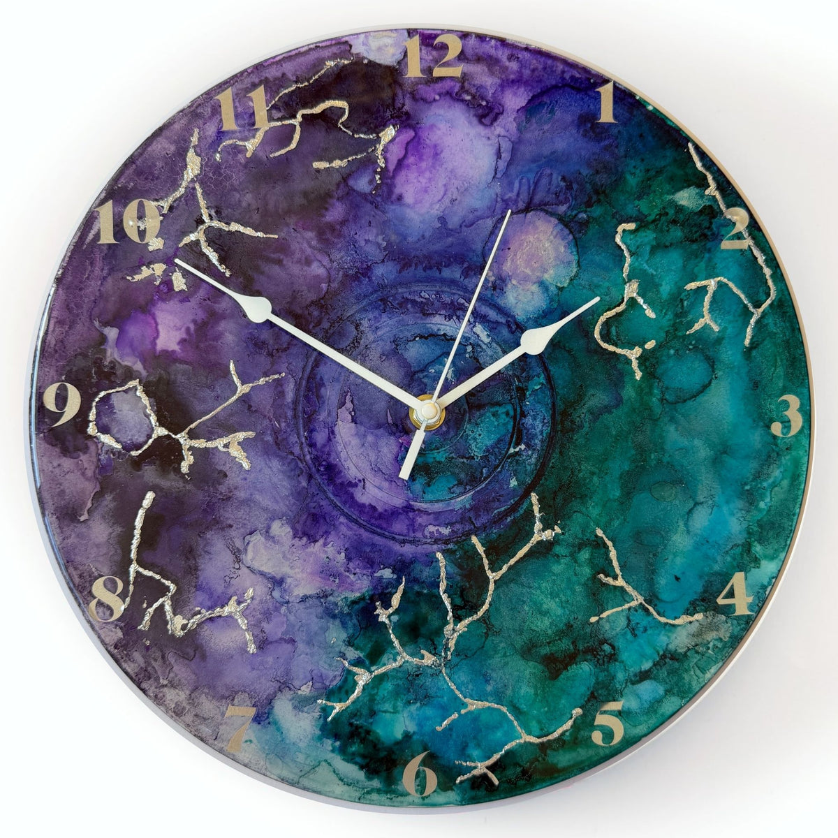 Round wall clock with a watercolour-style marbled face in purple, teal, and blue, with silver clock hands and silver Arabic numerals.