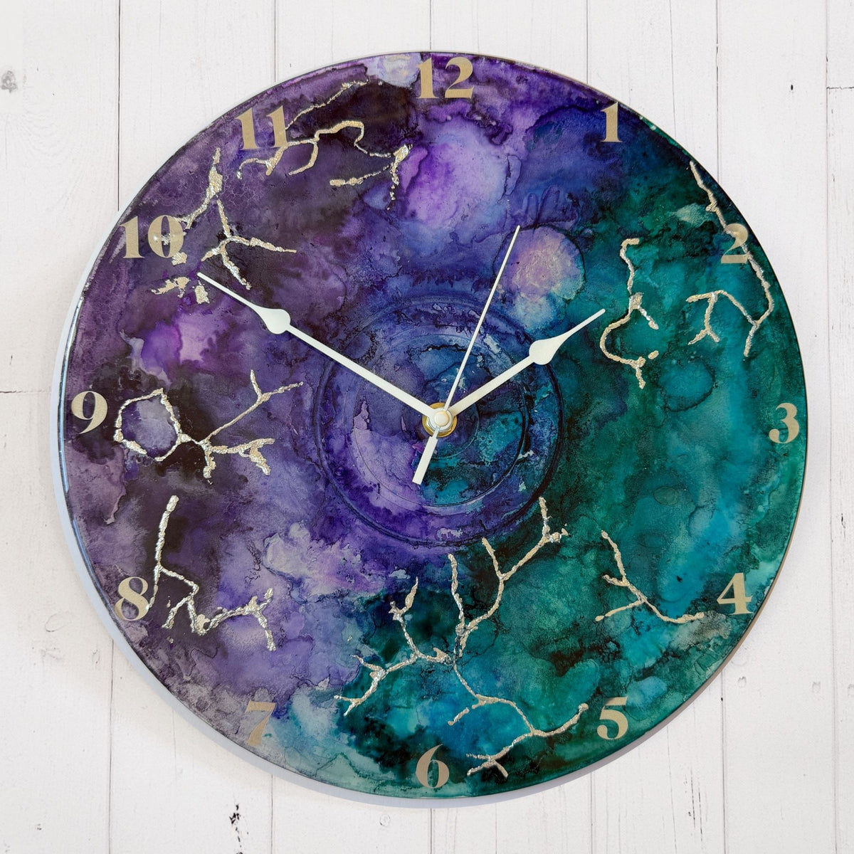 Round wall clock with a watercolour-style marbled face in purple, teal, and blue, with silver clock hands and silver Arabic numerals.