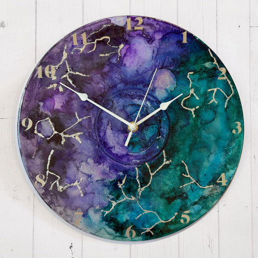 Round wall clock with a watercolour-style marbled face in purple, teal, and blue, with silver clock hands and silver Arabic numerals.