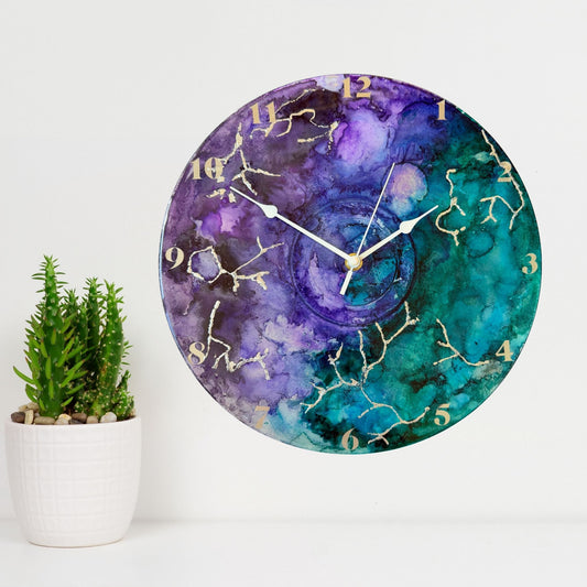Round wall clock with a watercolour-style marbled face in purple, teal, and blue, with silver clock hands and silver Arabic numerals.