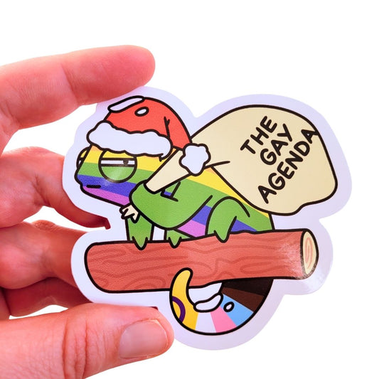 Sticker of a chmeleon holding a lbag with 'The Gay Agenda' text, held by a hand.